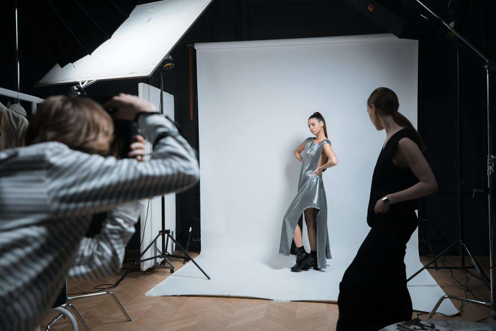 A working set in the UONE Brand Studio Brooklyn studio — two stylists pinning a garment on a model while a photographer reviews tethered captures on a calibrated monitor.